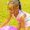 A young girl with twisted  hair wearing purple Bead Barrettes is playing with a large purple ball on a green grassy lawn. 