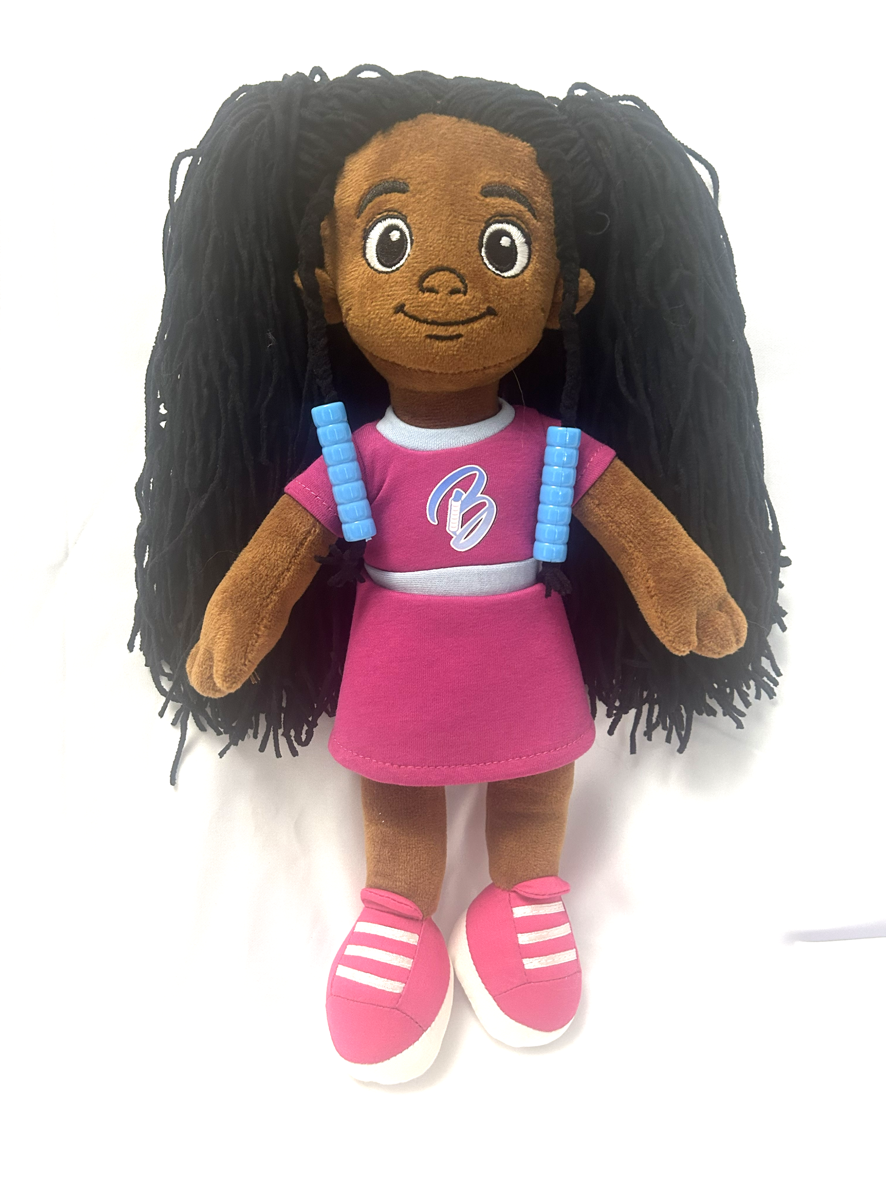 A plush Bead Barrettes  doll with medium brown skin, large cartoon eyes, and long black yarn hair. The doll is wearing two Bead Barrettes, a pink dress with a white collar, blue shoulder straps, and pink sneakers with white stripes, set against a plain white background.