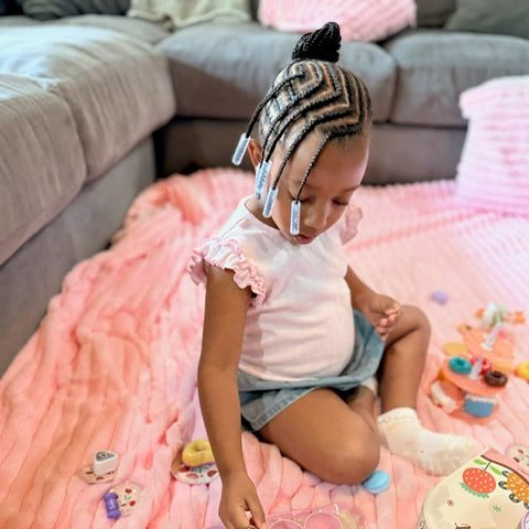 Child with braided hair wearing clear Bead Barrettes playing with a toy on a pink blanket in a living room.